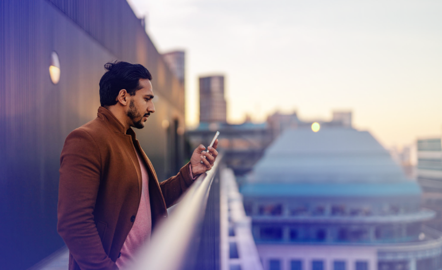 Person using a smartphone on a rooftop terrace overlooking a city. The image shows a modern urban environment with office buildings and soft evening lighting, representing connected, technology-driven work.